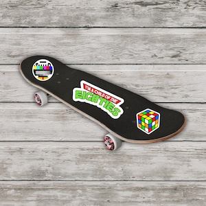 May include: Black skateboard with a retro theme. It features stickers of a TV test pattern, a Rubik's Cube, and the text "I'm a child of the Eighties" in green and white. The wheels are white with pink accents.