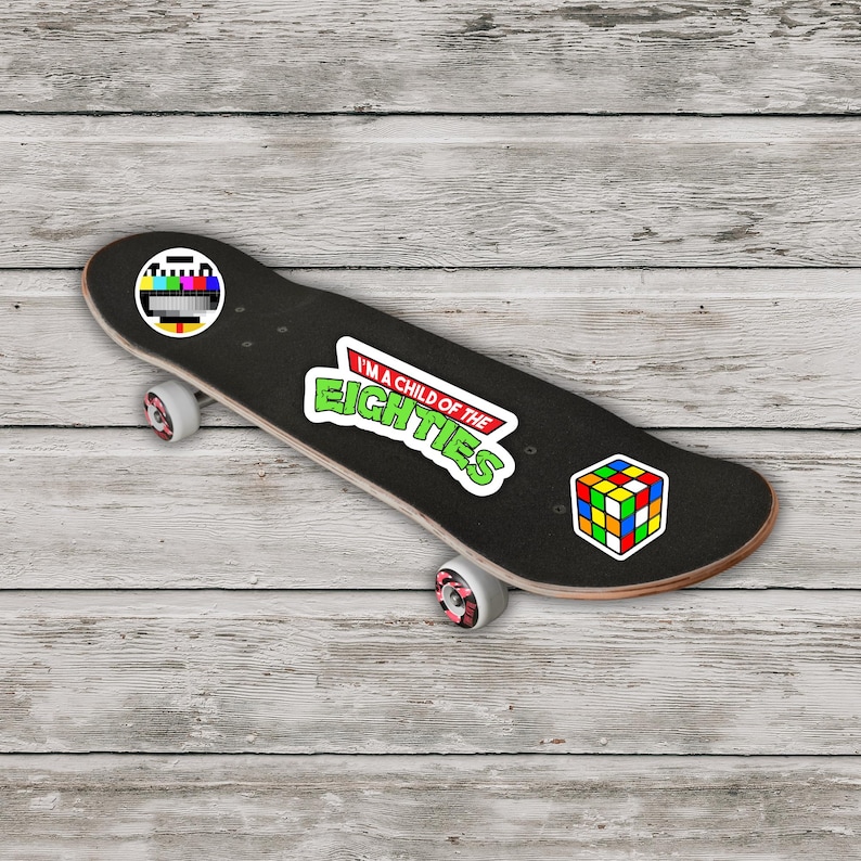 May include: Black skateboard with a retro theme. It features stickers of a TV test pattern, a Rubik's Cube, and the text "I'm a child of the Eighties" in green and white. The wheels are white with pink accents.
