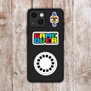 May include: Black phone case with multiple stickers. The stickers include a pixelated "GAME OVER" text, a white circular design with black details, and a cartoon burger with a blue and white border.