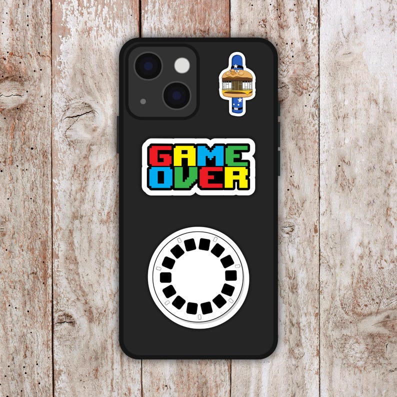 May include: Black phone case with multiple stickers. The stickers include a pixelated "GAME OVER" text, a white circular design with black details, and a cartoon burger with a blue and white border.