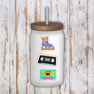 May include: White tumbler with a wooden lid and straw, adorned with stickers. Stickers include a colorful roller skate, a cassette tape labeled "BE KIND REWIND," and a 90s mixtape design.