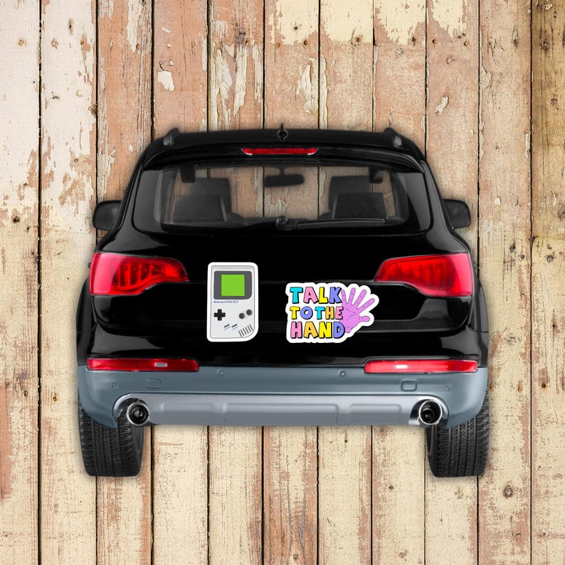 May include: Black car with a retro video game sticker and a sticker that says "Talk to the Hand". The video game sticker is white with a green screen. The hand sticker is multi-colored.