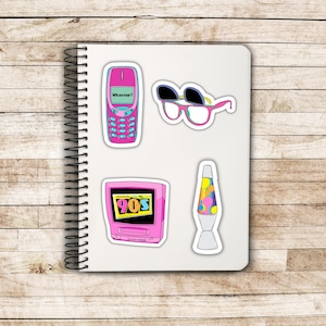 May include: A collection of colorful 90s-themed stickers on a white notebook. Includes a pink flip phone with "Whassup?" text, sunglasses, a pink TV with "90s" text, and a lava lamp.