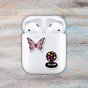 Puede incluir: Funda blanca para auriculares inal&aacute;mbricos con dos pegatinas: una mariposa rosa con purpurina y una bola de discoteca colorida. La funda est&aacute; abierta, revelando los auriculares en su interior. El fondo es una superficie de madera azul claro y gris desgastada.