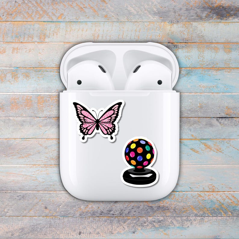 Puede incluir: Funda blanca para auriculares inal&aacute;mbricos con dos pegatinas: una mariposa rosa con purpurina y una bola de discoteca colorida. La funda est&aacute; abierta, revelando los auriculares en su interior. El fondo es una superficie de madera azul claro y gris desgastada.