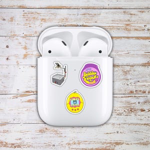 May include: White wireless earbuds in a charging case, adorned with three colorful stickers. The stickers feature a retro radio, a purple bubble tape design, and a yellow handheld game.