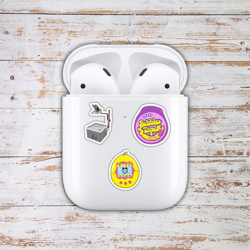 May include: White wireless earbuds in a charging case, adorned with three colorful stickers. The stickers feature a retro radio, a purple bubble tape design, and a yellow handheld game.