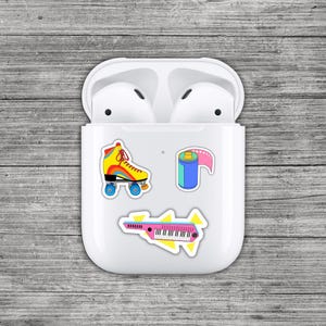 May include: White AirPods case with colorful stickers of a roller skate, film roll, and a synthesizer. The roller skate is yellow, red, and blue. The film roll is blue and pink. The synthesizer is pink and black.