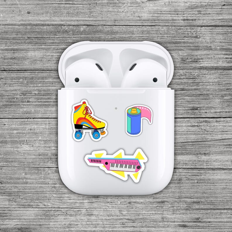 May include: White AirPods case with colorful stickers of a roller skate, film roll, and a synthesizer. The roller skate is yellow, red, and blue. The film roll is blue and pink. The synthesizer is pink and black.