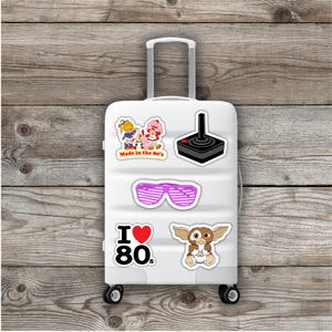 May include: A white suitcase decorated with retro stickers. Stickers include a joystick, pink shutter shades, a Gremlin, and a graphic that says "I ❤️ 80s". Other stickers say "Made in the 80's". The suitcase has a retractable handle and wheels.