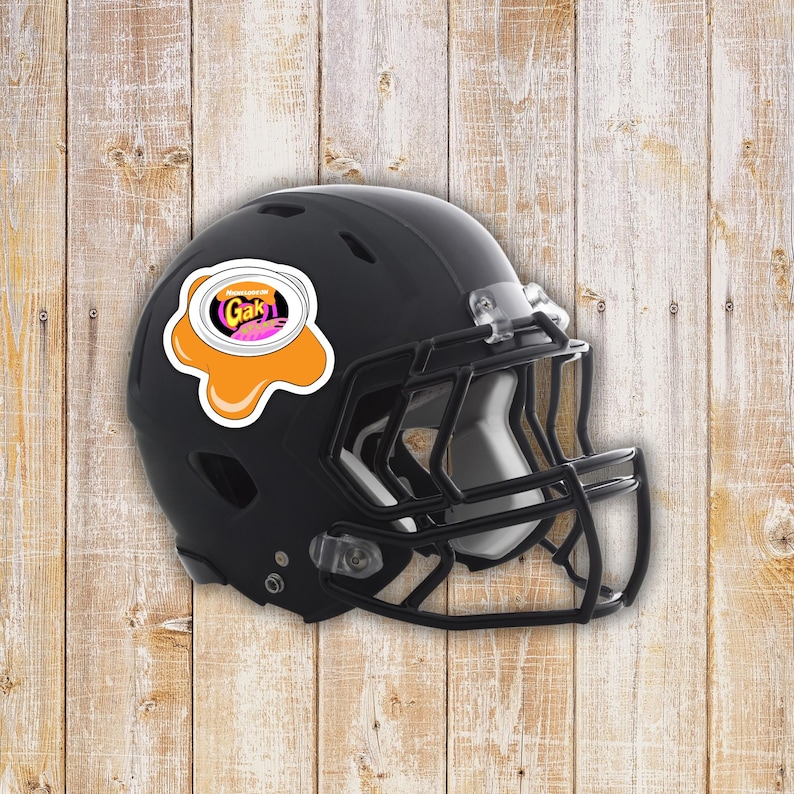 May include: Black football helmet with a sticker featuring the word "Gak" in a colorful design. The sticker is placed on the side of the helmet. The helmet has a black face mask and is set against a wooden background.
