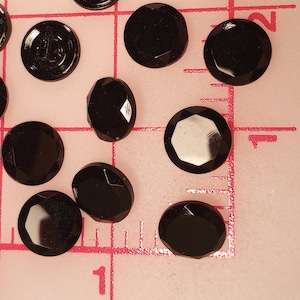 May include: A collection of black, faceted, round, and oval-shaped buttons. The buttons are scattered on a pink surface with a white grid.