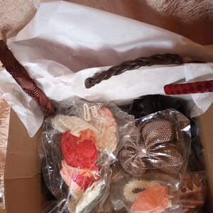 May include: A cardboard box filled with various hair accessories, including braided headbands in brown and red, and several packages of hair bows and flower accents in shades of pink, peach, and brown. The items are wrapped in clear plastic.