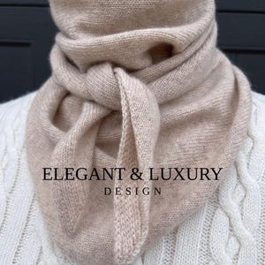 May include: A light beige knitted scarf with a knotted design. The scarf is made of a soft, textured material and is draped around the neck. The text "ELEGANT & LUXURY DESIGN" is printed on the scarf.