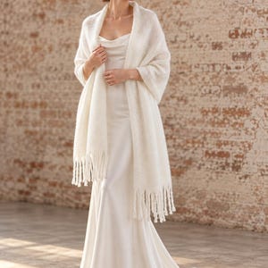 Puede incluir: Un vestido largo de satén color marfil sin mangas con escote tipo capucha y una ligera cola, combinado con un chal de punto suave y difuso en color blanco roto con largos flecos colgantes en los bordes inferiores, sobre los hombros y brazos.