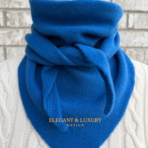 May include: A vibrant blue scarf with a triangular shape, tied at the front. The scarf is made of a soft, textured material and is draped over a white cable-knit sweater. The text "ELEGANT & LUXURY DESIGN" is printed on the scarf.