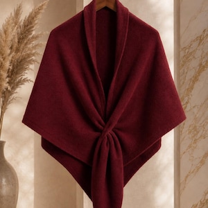 May include: A deep burgundy shawl with a textured knit finish, featuring a wide collar and a central knotted front design. The garment is draped symmetrically, creating a pointed hemline that hangs from a wooden hanger on a metal stand.
