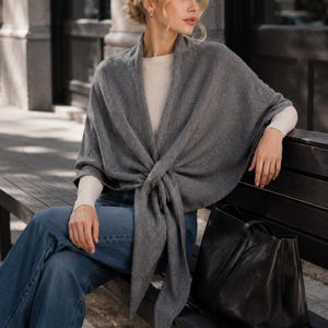 May include: A charcoal grey knit wrap shawl featuring a front tie closure, worn over a cream-coloured long-sleeve top and blue denim jeans, accompanied by a large black leather tote bag with dual handles and a structured rectangular base.