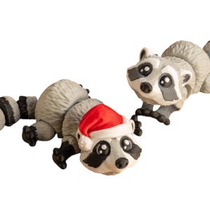 Könnte beinhalten: Zwei graue Waschbär-Figuren, eine mit einem roten Weihnachtsmann-Hut. Die Figuren haben schwarze Augen und Nasen und sind spielerisch posiert.