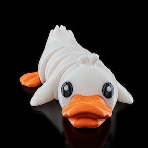 Könnte beinhalten: Eine weiße, 3D-gedruckte Ente mit orangefarbenem Schnabel und schwarzen Augen. Die Ente liegt auf dem Bauch und hat den Kopf zur Seite gedreht.