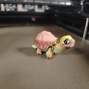 Könnte beinhalten: Eine 3D-gedruckte Schildkrötenfigur mit einem rosa-braunen Panzer, einem gelb-blauen Kopf und grünen Beinen. Die Schildkröte hat ein cartoonartiges Design mit großen Augen und einem lächelnden Gesichtsausdruck. Die Figur steht auf einer grauen Oberfläche.