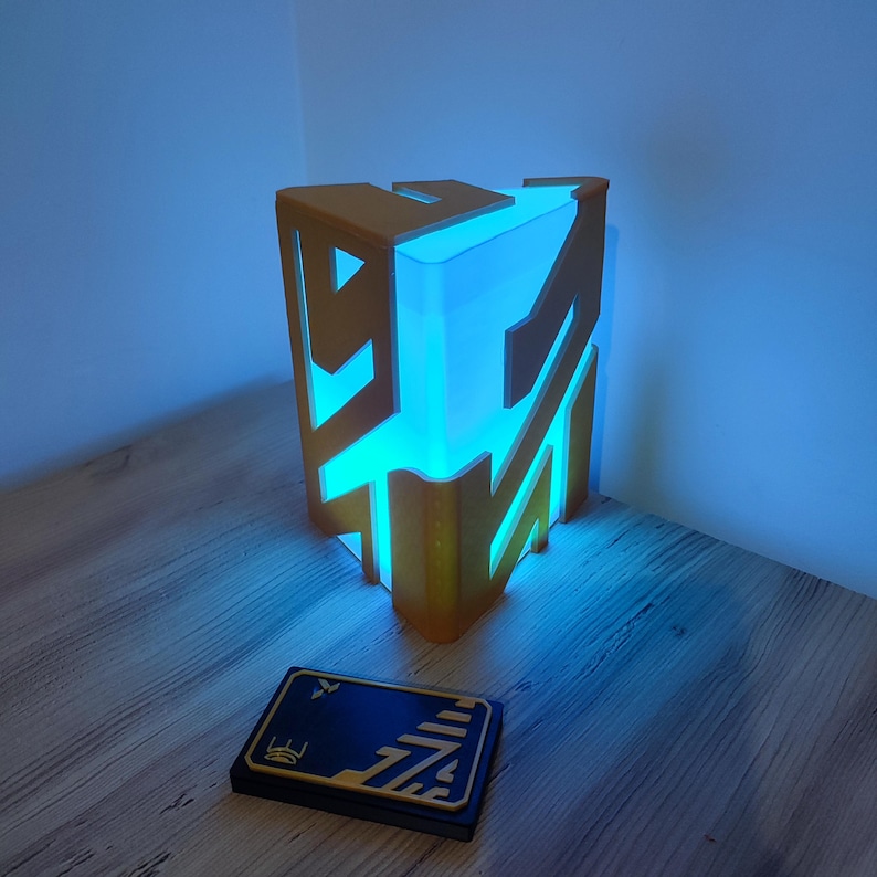 K&ouml;nnte beinhalten: Eine gold- und blaufarbene 3D-gedruckte Lampe in Form eines geometrischen W&uuml;rfels mit einem leuchtend blauen Licht im Inneren. Eine kleine schwarze Karte mit goldenen Akzenten befindet sich im Vordergrund.