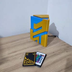 K&ouml;nnte beinhalten: Eine 3D-gedruckte geometrische Lampe in Blau und Gold mit einer Fernbedienung zum &Auml;ndern der Lichtfarbe. Die Fernbedienung hat ein schwarzes und gelbes Design mit den Buchstaben "DE" darauf.