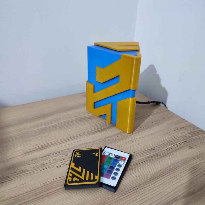 K&ouml;nnte beinhalten: Eine 3D-gedruckte geometrische Lampe in Blau und Gold mit einer Fernbedienung zum &Auml;ndern der Lichtfarbe. Die Fernbedienung hat ein schwarzes und gelbes Design mit den Buchstaben "DE" darauf.
