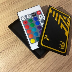 K&ouml;nnte beinhalten: Eine schwarze Fernbedienung mit einer Vielzahl von farbigen Kn&ouml;pfen, darunter rot, gr&uuml;n, blau, gelb und wei&szlig;. Die Fernbedienung hat die Aufschriften "OFF", "ON", "W" und "FLASH" aufgedruckt. Die Fernbedienung liegt auf einem schwarzen Geh&auml;use mit einem gelben und schwarzen Design.