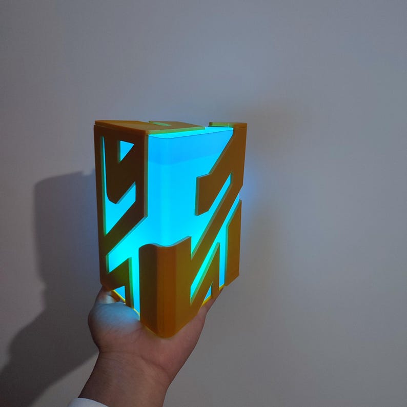 K&ouml;nnte beinhalten: Eine 3D-gedruckte, goldfarbene Box mit einem leuchtend blauen Licht im Inneren. Die Box hat ein geometrisches Design mit einem Muster aus Linien und Formen.