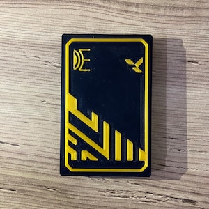 K&ouml;nnte beinhalten: Eine schwarze Karte mit gelbem Rand und einem gelben geometrischen Muster. Die Karte hat ein gelbes Symbol in der oberen rechten Ecke und den Buchstaben "E" in der oberen linken Ecke.