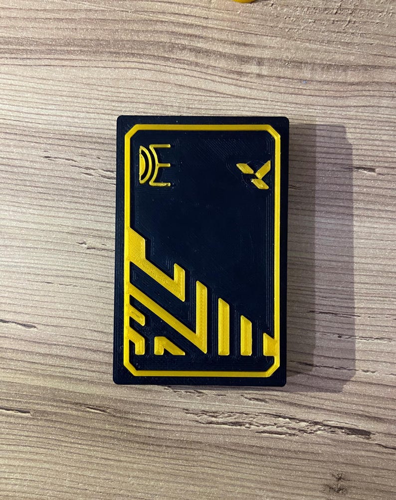 K&ouml;nnte beinhalten: Eine schwarze Karte mit gelbem Rand und einem gelben geometrischen Muster. Die Karte hat ein gelbes Symbol in der oberen rechten Ecke und den Buchstaben "E" in der oberen linken Ecke.