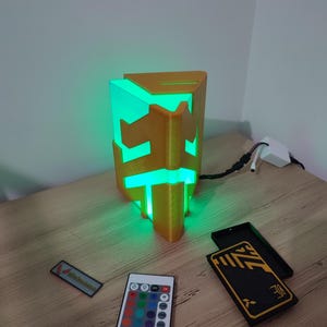 K&ouml;nnte beinhalten: Eine 3D-gedruckte, goldfarbene Lampe in Form eines geometrischen Prismas mit einem gr&uuml;nen Licht im Inneren. Die Lampe steht auf einem Holztisch mit einer Fernbedienung und zwei schwarzen Boxen mit goldenen Akzenten.
