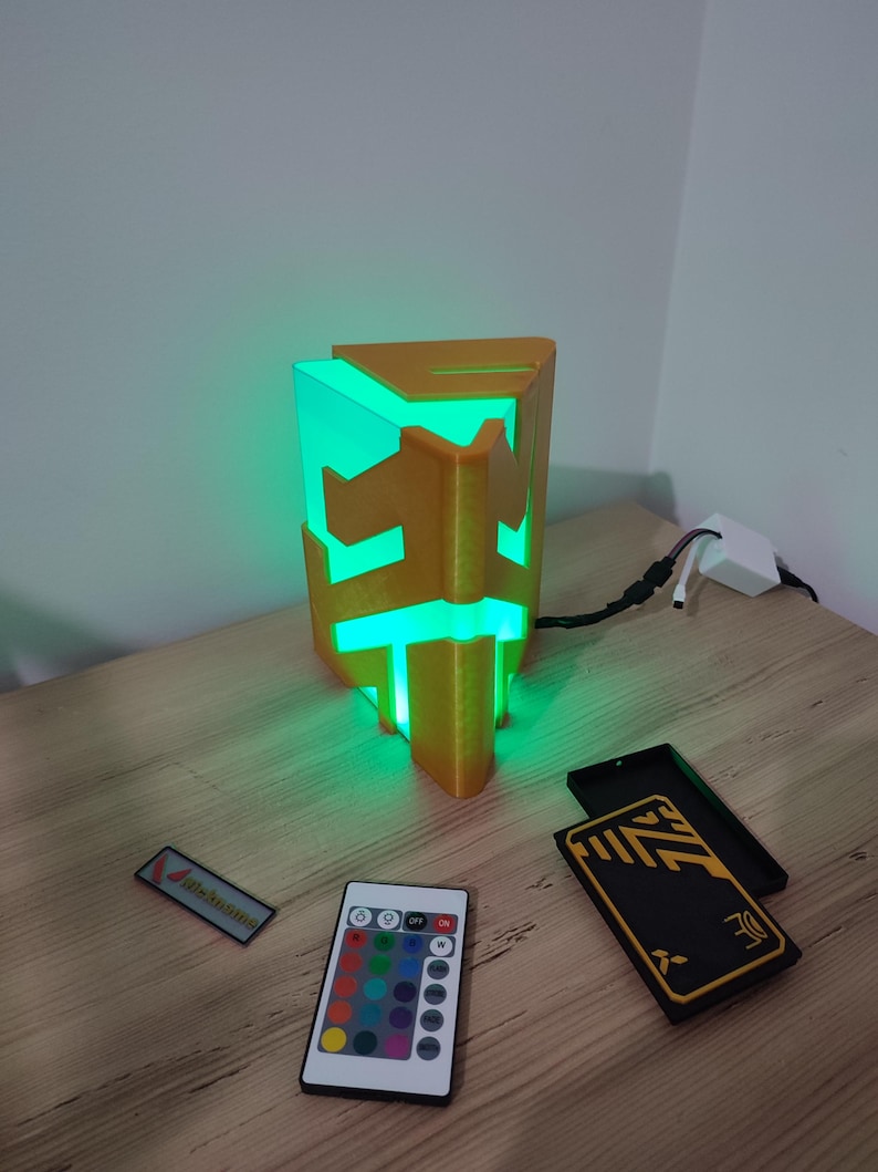 K&ouml;nnte beinhalten: Eine 3D-gedruckte, goldfarbene Lampe in Form eines geometrischen Prismas mit einem gr&uuml;nen Licht im Inneren. Die Lampe steht auf einem Holztisch mit einer Fernbedienung und zwei schwarzen Boxen mit goldenen Akzenten.