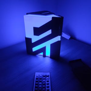 K&ouml;nnte beinhalten: Ein blaues Licht mit einem schwarzen Rahmen und einer Fernbedienung. Das Licht ist in Form eines Buchstabens "F" gestaltet.