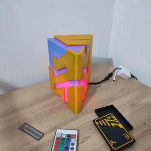 K&ouml;nnte beinhalten: Eine 3D-gedruckte geometrische Lampe mit gelbem und blauem Design. Die Lampe wird mit einem mehrfarbigen LED-Licht beleuchtet und wird mit einer Fernbedienung geliefert.