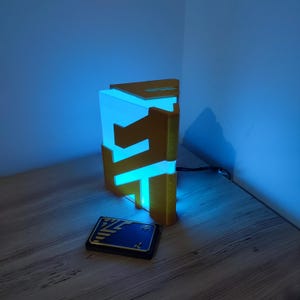 May include: A 3D printed lamp shaped like a stylised letter "E" with a blue light shining through the translucent plastic. The lamp is sitting on a wooden surface next to a small blue and black card with a stylised "E" on it.