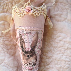 May include: A pink fabric cone with lace trim and a pink rose. The cone has a printed image of a rabbit with pink roses and the text "J. Blondeel & Cie. 38 Rue des Jeunesses 33."