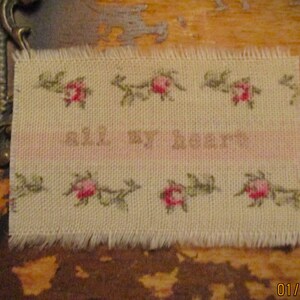 Peut inclure: Un échantillon de tissu beige avec une bande rose et des fleurs brodées ainsi que le texte "All My Heart".