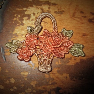 Può includere: Un delicato cestino ricamato pieno di fiori arancioni e dorati. Il cestino è fatto di pizzo e ha un manico. I fiori sono disposti in un bouquet e hanno un aspetto vintage.