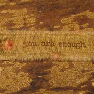 May include: A tan burlap ribbon with frayed edges, featuring the phrase "you are enough" in brown lettering. The ribbon is adorned with small pink rose illustrations. The background is a textured, brown surface.