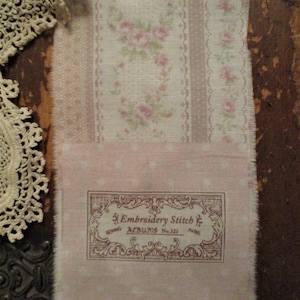 Könnte beinhalten: Ein rosa-weißes Stoffstück mit einem braunen Etikett, auf dem "Embroidery Stitch Sewing Albums No. 325 Paris" steht.
