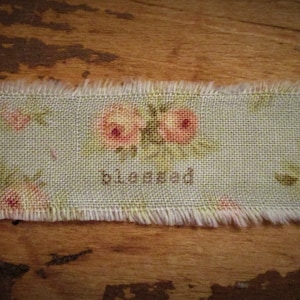 May include: A light blue fabric with a frayed edge features a floral pattern with pink flowers and the word "blessed" printed in brown.