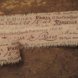 May include: A section of vintage-style fabric with frayed edges, featuring faded text in dark brown ink. The text includes the words "FABRIQUE," "HUILES," "PARIS," and "MARSEILLE." The fabric has a light brown background.