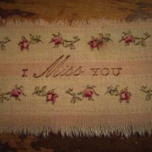 May include: A small, rectangular piece of fabric with a frayed edge. It is embroidered with pink flowers and the words "I Miss You".