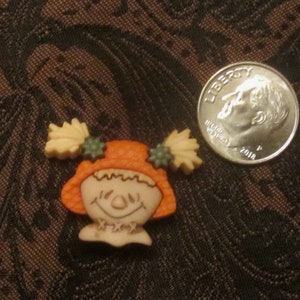 Può includere: Un piccolo bottone arancione e beige con un viso di spaventapasseri sorridente e foglie verdi e beige sui lati.