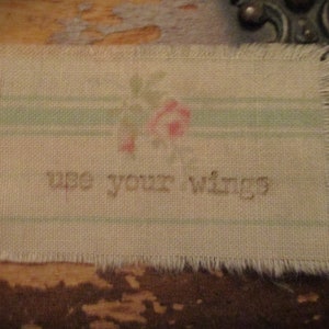 May include: A rectangular fabric patch with frayed edges. The patch features a faded floral design and the words "use your wings" in a simple font. Light green stripes run horizontally across the fabric.