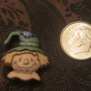 Può includere: Una piccola statuina di spaventapasseri in ceramica con un cappello verde, capelli castani e un viso amichevole. La statuina è circa delle dimensioni di una moneta da dieci centesimi di euro.