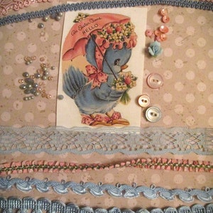 Könnte beinhalten: Eine Vintage-Osterkarte mit einem blauen Vogel, der einen Regenschirm mit dem Text "An Easter Time Hello" hält. Die Karte ist von blauem und rosa Spitzenbesatz, Perlen und Knöpfen umgeben.