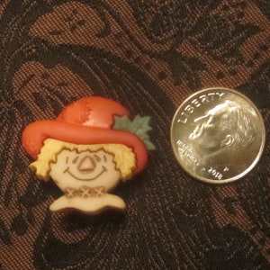 Può includere: Una piccola statuina di spaventapasseri di colore crema con un cappello rosso e foglie verdi. Lo spaventapasseri ha un viso sorridente e un motivo a croce cucito sul petto.